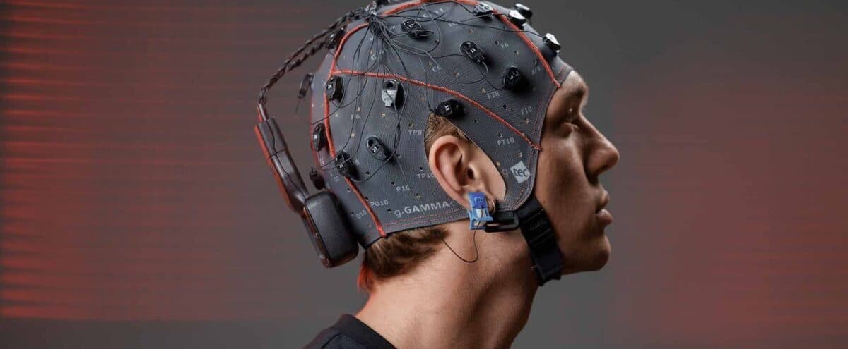 Profilden bir adam; başında elektrotlu gri bir EEG başlığı, başlığın arkasında kablosuz bir amplifikatör ve kulağında mavi bir referans klipsi